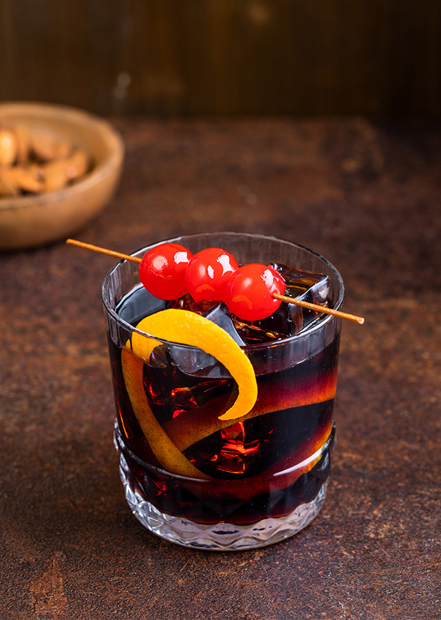 Glas mit dunklem Cocktail, garniert mit einer Zitronenzeste und drei roten Kirschen auf einem Holzspieß, im Hintergrund eine Schale mit Nüssen