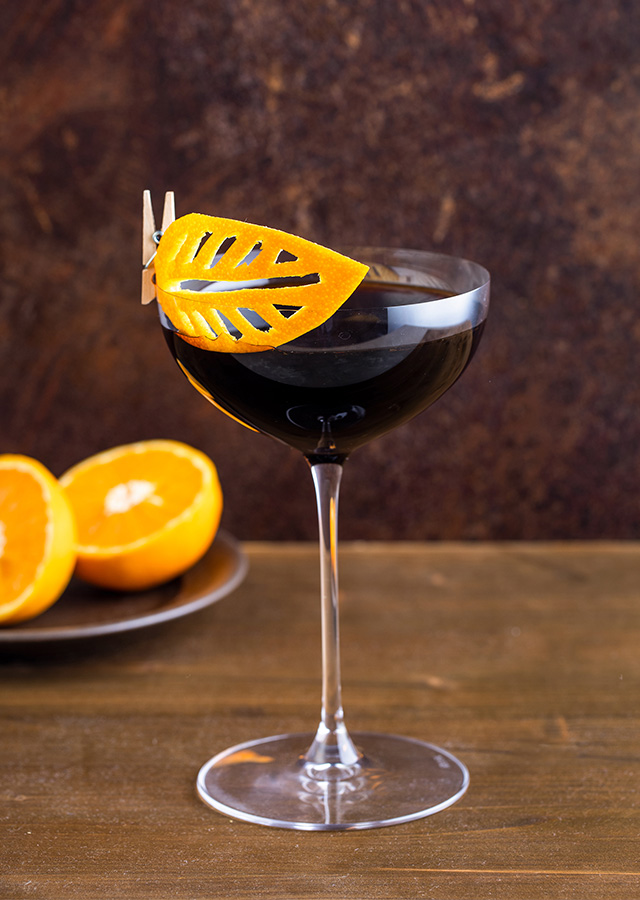 Cocktailglas mit dunklem Getränk und orangefarbener Dekoration am Glasrand vor Teller mit Orangenhälften auf Holztisch