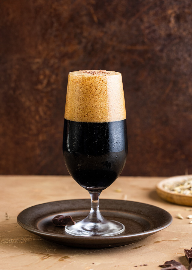 Glas mit dunklem Bier und cremigem Schaum auf einem dunklen Untersetzer vor braunem Hintergrund mit Schokoladenstücken und Schalen mit Körnern