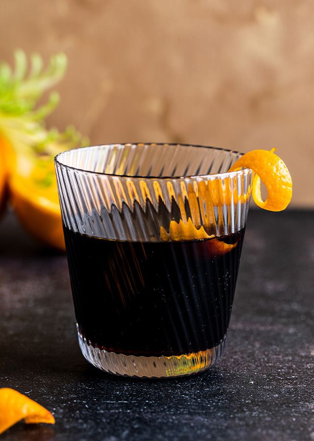 Glas mit dunklem Cocktail und Zitronenzeste auf dunklem Untergrund mit Zitronen im Hintergrund