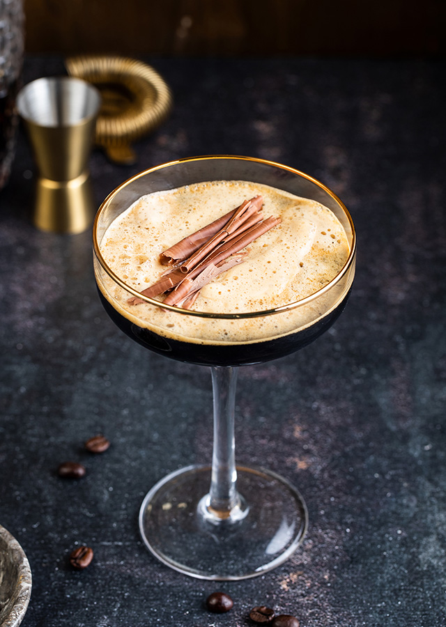 Cocktailglas mit schaumigem Espresso Martini und Zimtstangen auf dunklem Untergrund mit Kaffeebohnen und Messbecher im Hintergrund