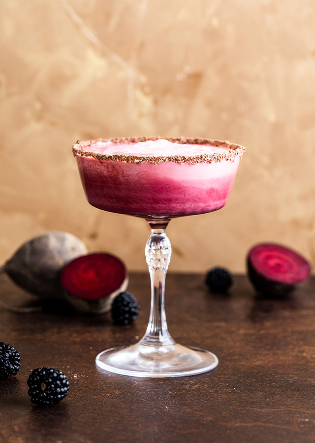 Cocktailglas mit rosa Schaum und Zuckerrand auf dunklem Untergrund umgeben von Brombeeren und halbierten Rote-Bete-Knollen