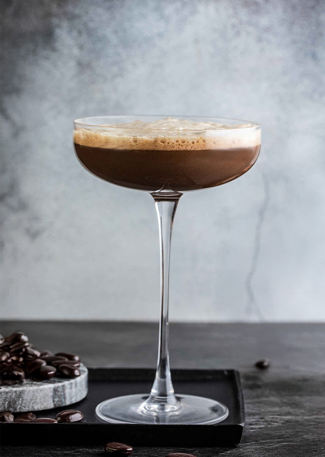 Cocktailglas mit Espresso-Martini auf dunklem Untergrund neben Kaffeebohnen und Schokoladenstücken