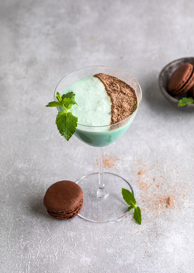 Cocktailglas mit grünem Getränk, mit Kakaopulver und Minzblatt garniert, daneben zwei braune Macarons auf grauem Untergrund