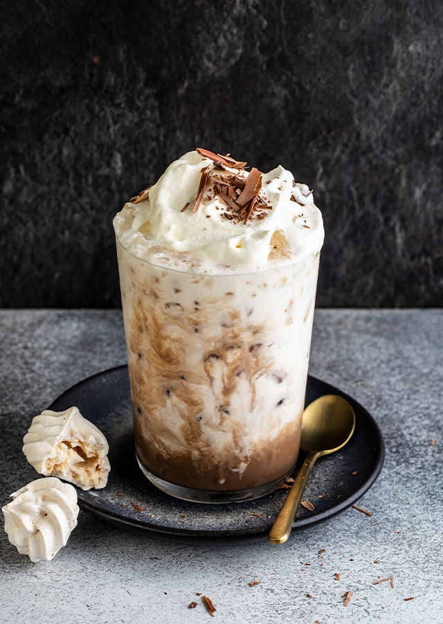 Glas mit Eiskaffeegetränk, Sahnehaube und Schokoraspeln auf dunklem Unterteller mit goldfarbenem Löffel und zwei kleinen Baiserstücken auf grauem Untergrund