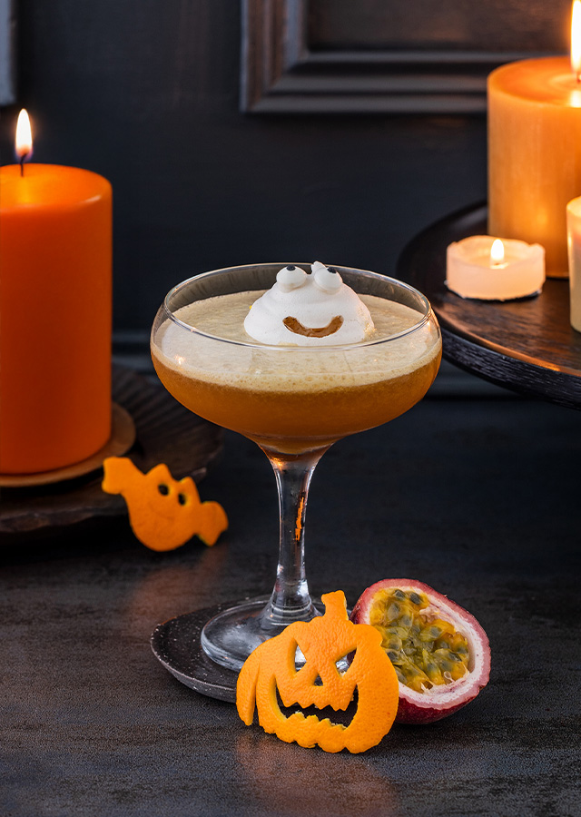 Cocktailglas mit Schaumhaube und lachendem Kürbis aus Orangenschale daneben, umgeben von brennenden Kerzen und einer halbierten Passionsfrucht auf dunklem Untergrund