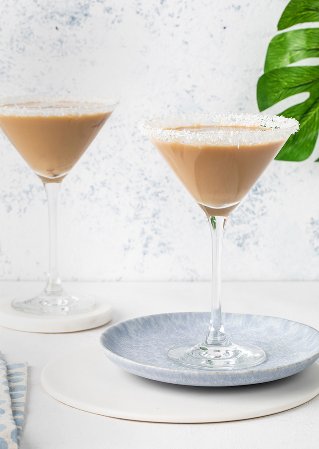 Zwei Martini-Gläser mit cremigem Kokos-Cocktail auf hellem Untergrund, eines auf einem grauen Teller, daneben ein grünes Blatt und ein gestreiftes Tuch