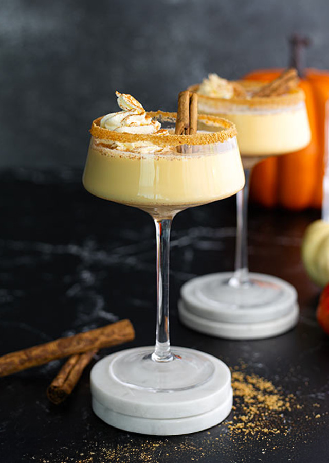 Zwei Cocktails mit Schlagsahne und Zimt auf Marmoruntersetzern vor Kürbissen und Glasflasche auf dunklem Untergrund