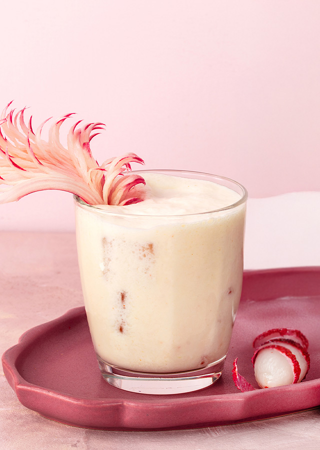 Glas mit cremigem Cocktail auf rosa Tablett neben zwei kleinen roten Kugeln und rosa Blume auf hellem Untergrund