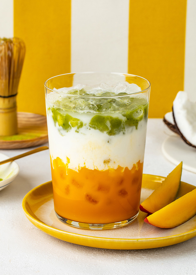 Glas mit geschichteter Mango-Matcha-Latte auf gelbem Teller mit Mangoscheiben daneben und halbierter Kokosnuss im Hintergrund