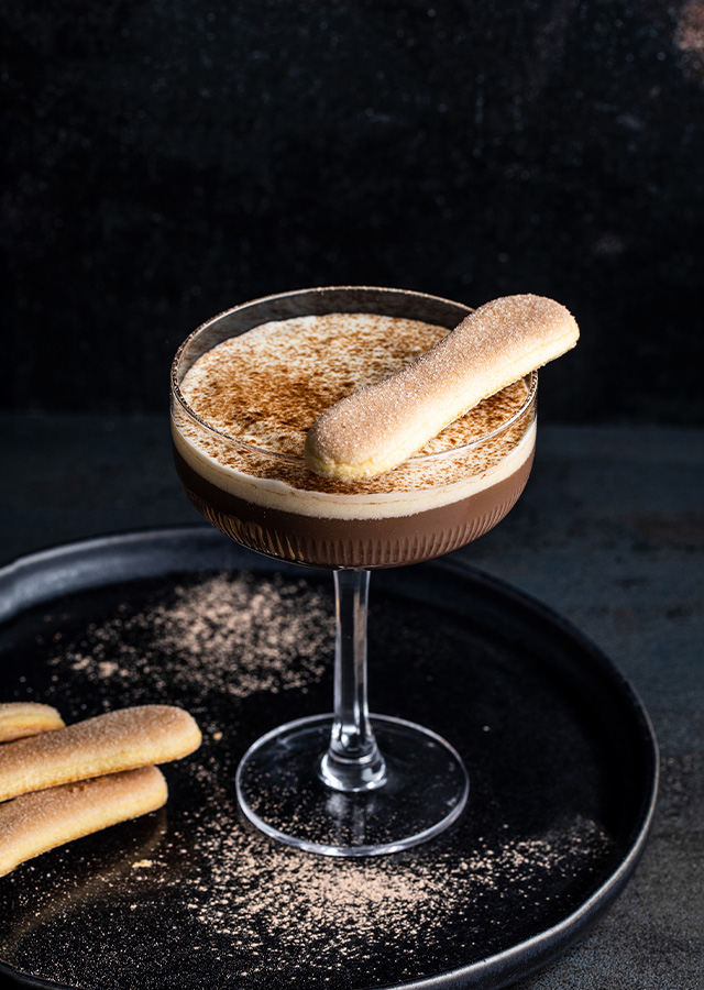 Cocktailglas mit Tiramisu-Martini und Löffelbiskuit auf schwarzem Teller vor dunklem HintergrundCocktailglas mit Tiramisu-Martini und Löffelbiskuit auf schwarzem Teller vor dunklem Hintergrund