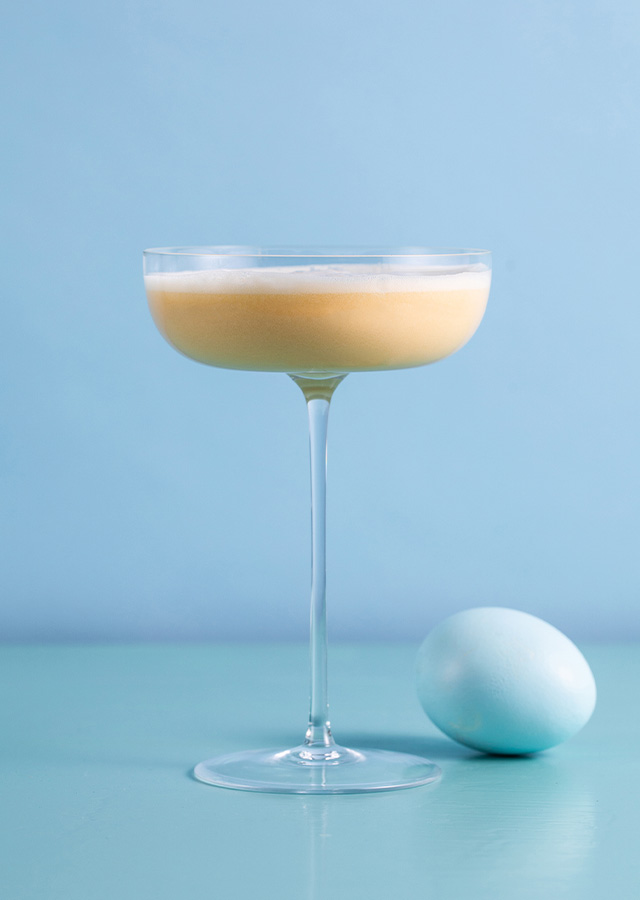 Cocktailglas mit cremigem hellgelbem Getränk neben einem weißen Ei auf hellblauem Untergrund vor hellblauem Hintergrund