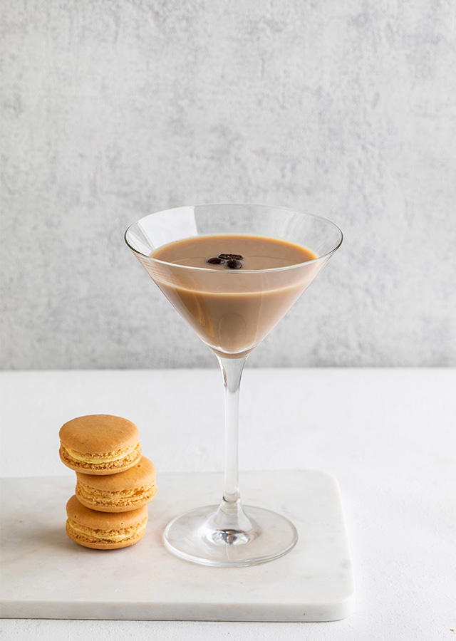 Martiniglas mit hellbraunem Espresso-Martini und drei Kaffeebohnen darauf neben zwei gestapelten hellbraunen Macarons auf weißem Marmorbrett vor hellgrauem Hintergrund