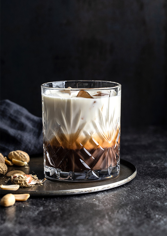 Glas mit geschichteter White-Russian-Cocktail auf dunklem Untergrund mit Nüssen und Stoffserviette