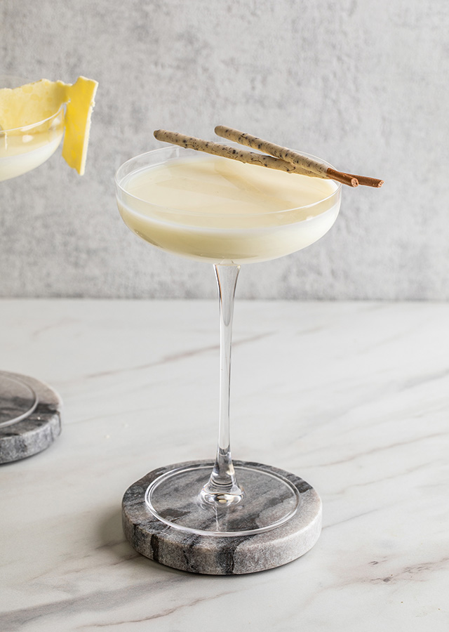 Zwei Cocktails in Martinigläsern mit weißer Schaumkrone auf Marmoruntersetzern, einer mit Zitronenscheibe, der andere mit zwei Zimtstangen garniert