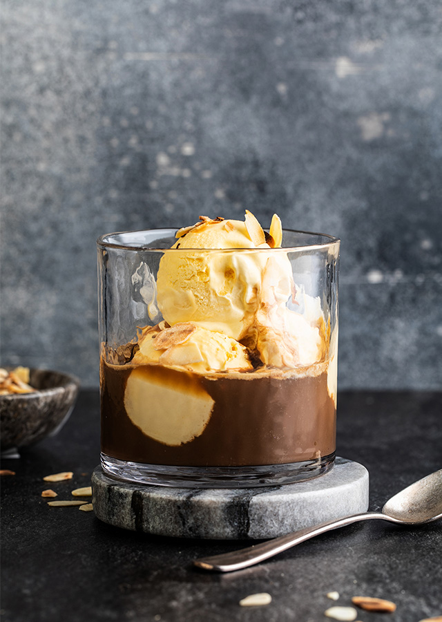 Glas mit Affogato bestehend aus Vanilleeis und Espresso auf grauem Marmoruntersetzer mit Löffel und Schale im Hintergrund