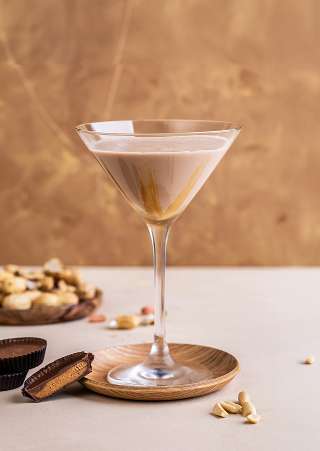 Nahaufnahme eines Martini-Glases mit cremigem Getränk und Erdnussbutter, daneben Schalen mit Erdnussbutter-Pralinen und Erdnüssen auf hellem Untergrund