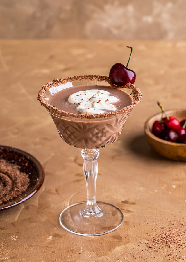 Cocktailglas mit Schokoladenmousse, Sahnehaube und Kirsche auf beigem Untergrund neben Schale mit Kirschen und Teller mit Kakaopulver