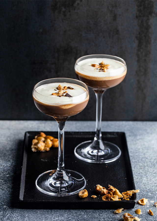 Zwei Cocktailgläser mit schaumiger Kaffee-Cocktail-Mischung auf schwarzem Tablett mit Kaffeebohnen und Kaffeebohnenstückchen auf grauem Untergrund