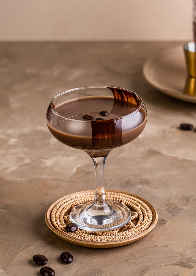 Cocktailglas mit dunkelbraunem Getränk, verziert mit Kaffeebohnen und Schokoladenstreifen, auf rundem Untersetzer