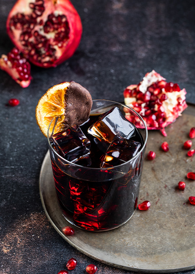 Glas mit dunklem Cocktail, Eiswürfeln und getrockneter Orangenscheibe auf grauem Teller mit Granatapfelkernen und aufgebrochenem Granatapfel im Hintergrund