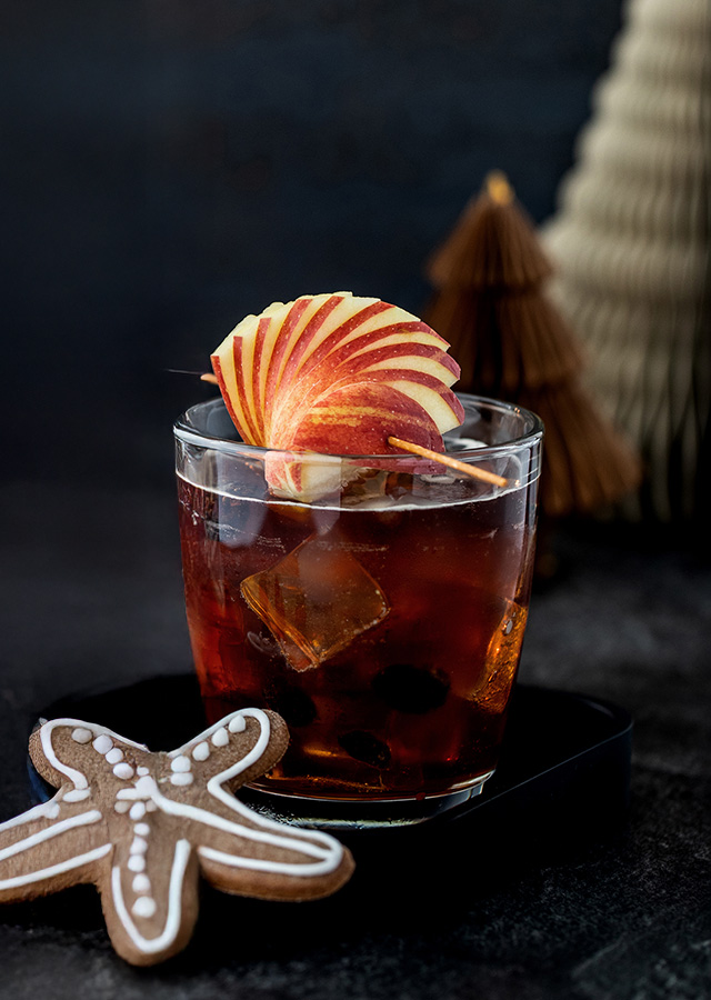 Glas mit dunklem Getränk, Eiswürfeln und Apfelscheiben als Garnitur neben sternförmigem Lebkuchen auf dunklem Untergrund