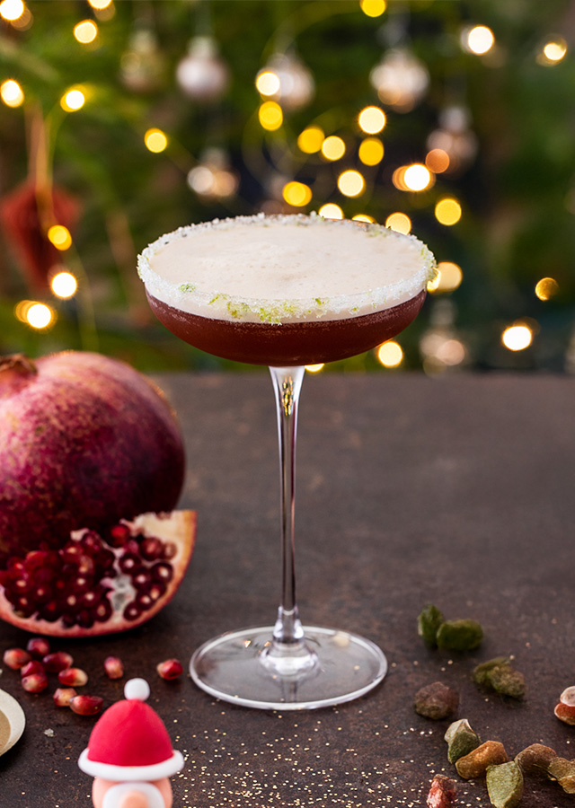 Cocktailglas mit schaumiger weißer Oberfläche auf dunklem Untergrund, im Hintergrund Granatapfel und kleine Figuren mit Weihnachtsmützen