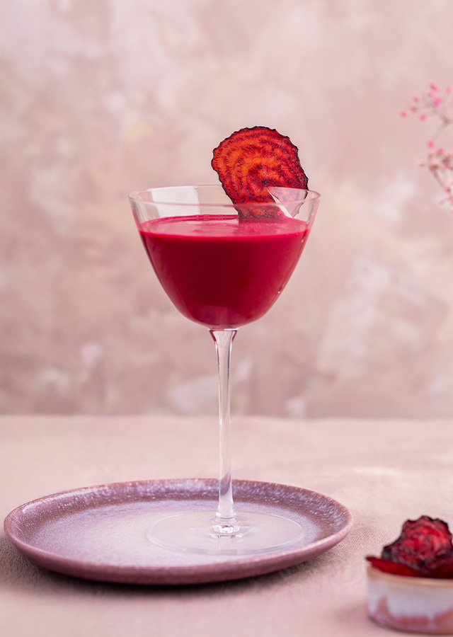 Cocktailglas mit dunkelrotem Getränk und getrockneter Fruchtscheibe auf einem Teller daneben ein kleines Glas mit weiteren getrockneten Fruchtscheiben und im Hintergrund eine Vase mit rosa Blumen