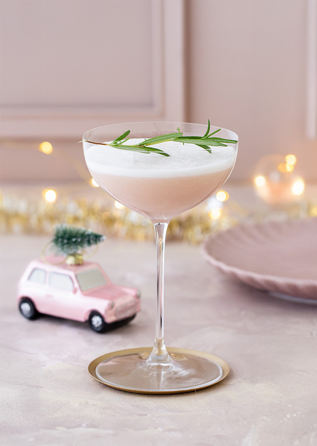 A cocktail glass containing a white drink and sprigs of rosemary on a table with a pink plate and a small toy car with a Christmas tree on its roof