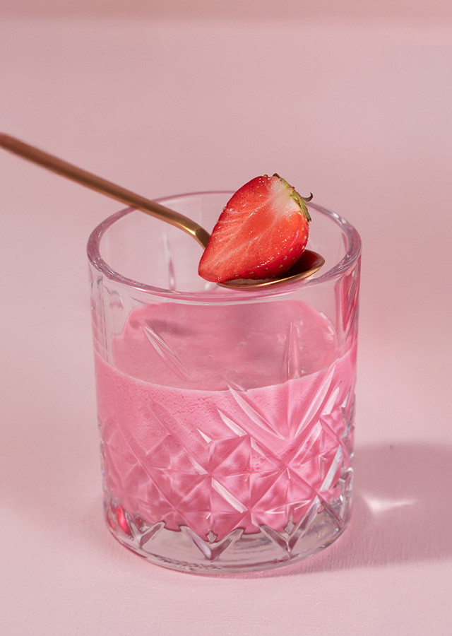 Kristallglas mit rosa Cocktail und halbierter Erdbeere auf Glasrand vor rosa Hintergrund