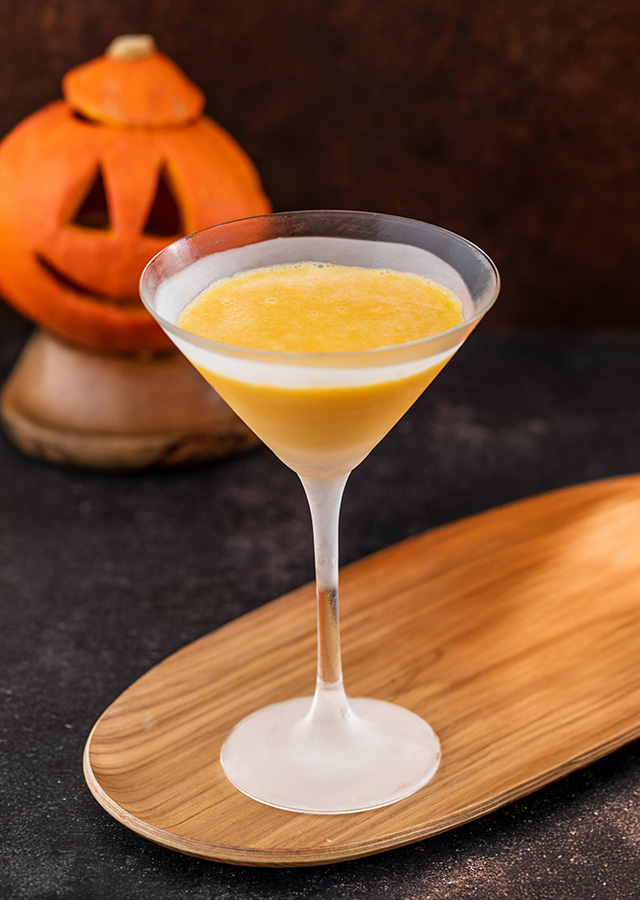 Cocktailglas mit gelb-orangefarbener Flüssigkeit auf einem länglichen Holztablett vor einem geschnitzten Kürbisgesicht auf dunklem Untergrund