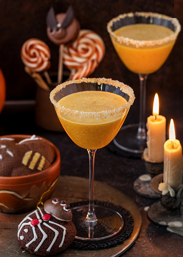 Zwei mit Zuckerrand verzierte Cocktailgläser mit orangefarbenem Getränk, daneben Halloween-Kürbis, Kerzen und Schokoladenkekse mit Spinnennetz-Dekor