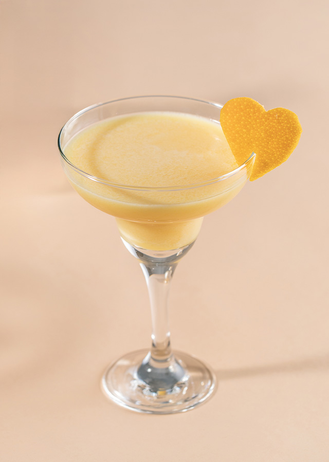 Cocktailglas mit hellgelbem Getränk und herzförmiger Orangenschale am Glasrand vor beigem Hintergrund