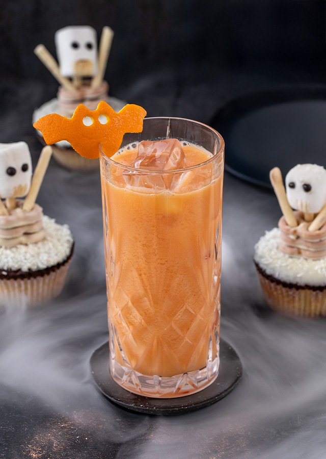 Glas mit orangefarbenem Getränk und Eiswürfeln auf schwarzem Untergrund umgeben von drei Cupcakes mit Skelett- und Fledermaus-Dekorationen und Nebeleffekt