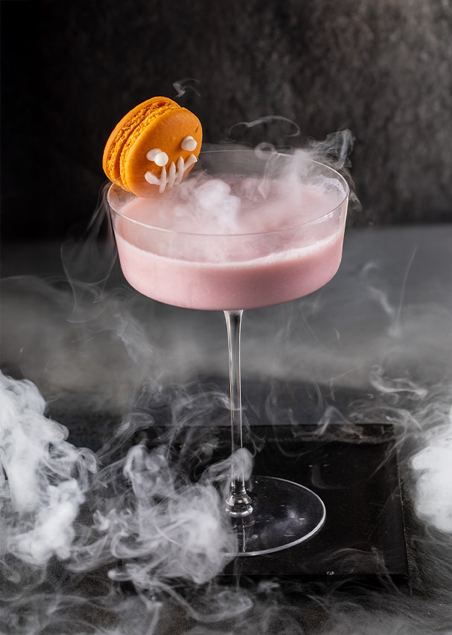 Cocktailglas mit rosa Getränk und orangefarbenem Keks mit Vampirzähnen auf Glasrand und schwarzem Tablett mit Nebeleffekt