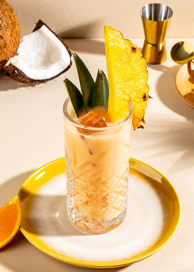 Glas mit hellbraunem Cocktail, garniert mit Ananasstück und grünen Blättern, auf weißem Teller mit gelbem Rand, daneben Orangenscheibe und halbierte Kokosnuss sowie goldfarbene Schale mit Eiswürfeln