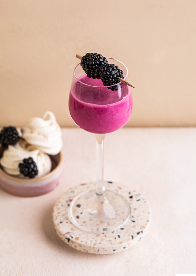 Glas mit pinkem Getränk und Brombeeren auf einem runden Steinuntersetzer neben zwei kleinen weißen Desserts mit Brombeeren auf hellem Untergrund
