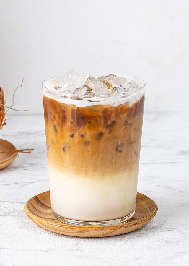 Glas mit Eiskaffee auf Holzuntersetzer vor halbierter Kokosnuss und Ananas auf weißer Marmoroberfläche
