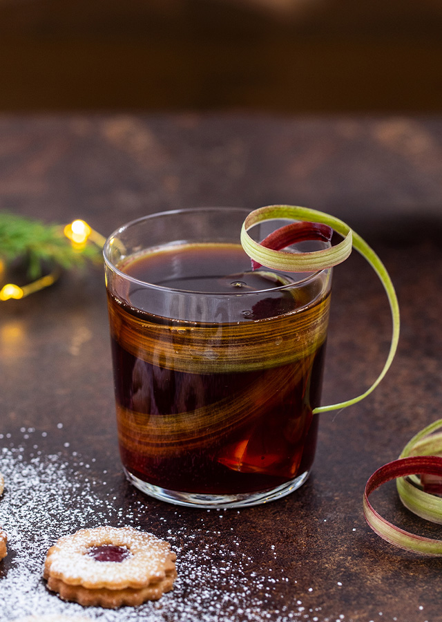 Glas mit dunklem Getränk und einer Spirale aus grün-rotem Gemüse oder Obst als Garnitur neben mehreren mit Puderzucker bestäubten Linzer Plätzchen auf dunklem Untergrund