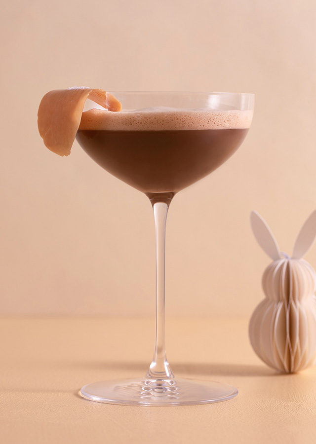 Cocktailglas mit braunem Getränk und Orangenzeste auf hellem Untergrund neben einer kleinen Papierfigur in Hasenform
