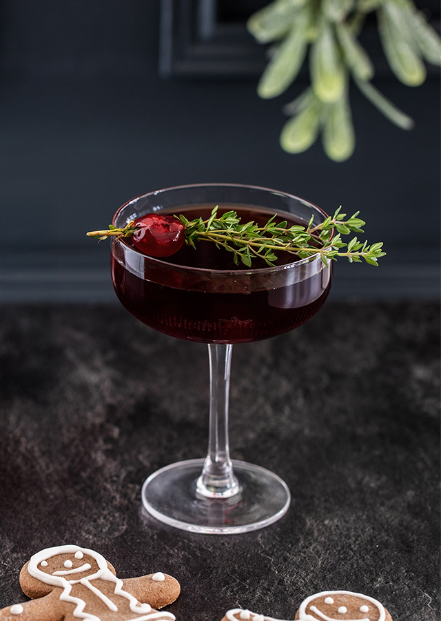 Cocktailglas mit dunklem Getränk, garniert mit einem Zweig Thymian und einer roten Beere