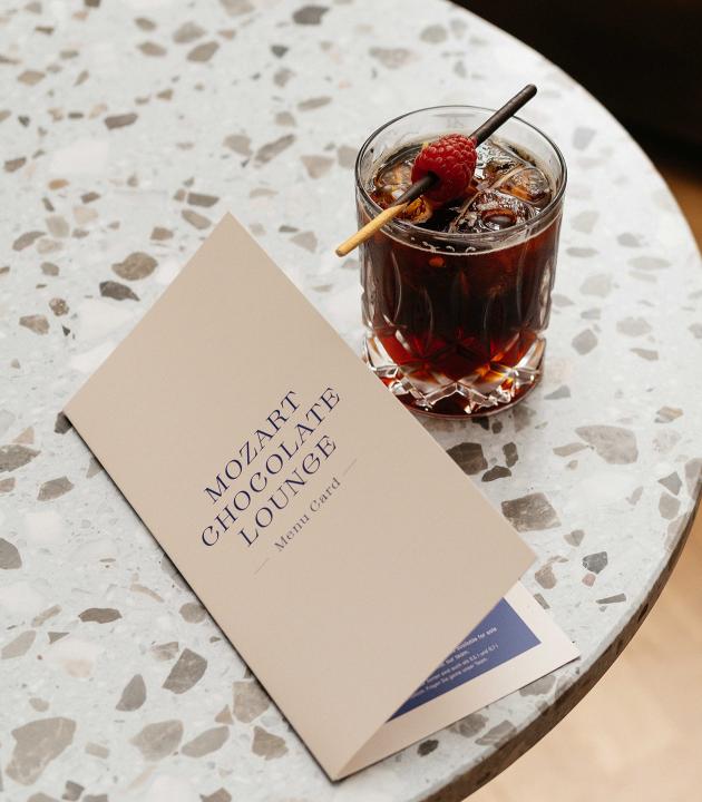 Ein Cocktail mit einer Himbeere und einem Holzspieß steht neben einem geöffneten Menü der 'Mozart Chocolate Lounge' auf einem Tisch aus Marmor.