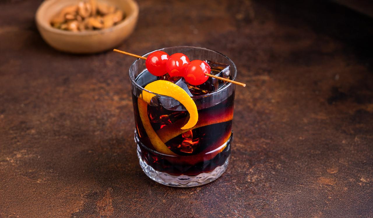 Glas mit dunklem Cocktail, garniert mit einer Zitronenzeste und drei roten Kirschen auf einem Holzspieß, im Hintergrund eine Schale mit Nüssen