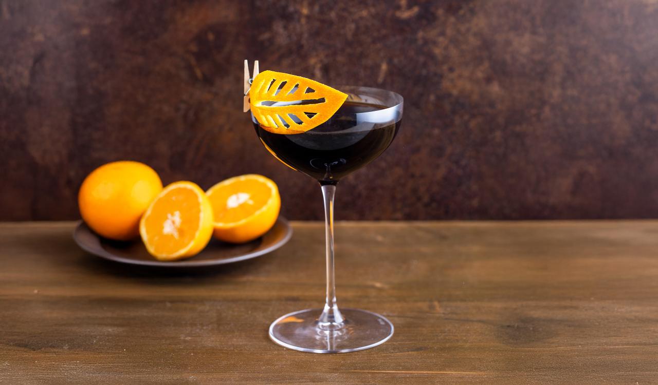 Cocktailglas mit dunklem Getränk und orangefarbener Dekoration am Glasrand vor Teller mit Orangenhälften auf Holztisch