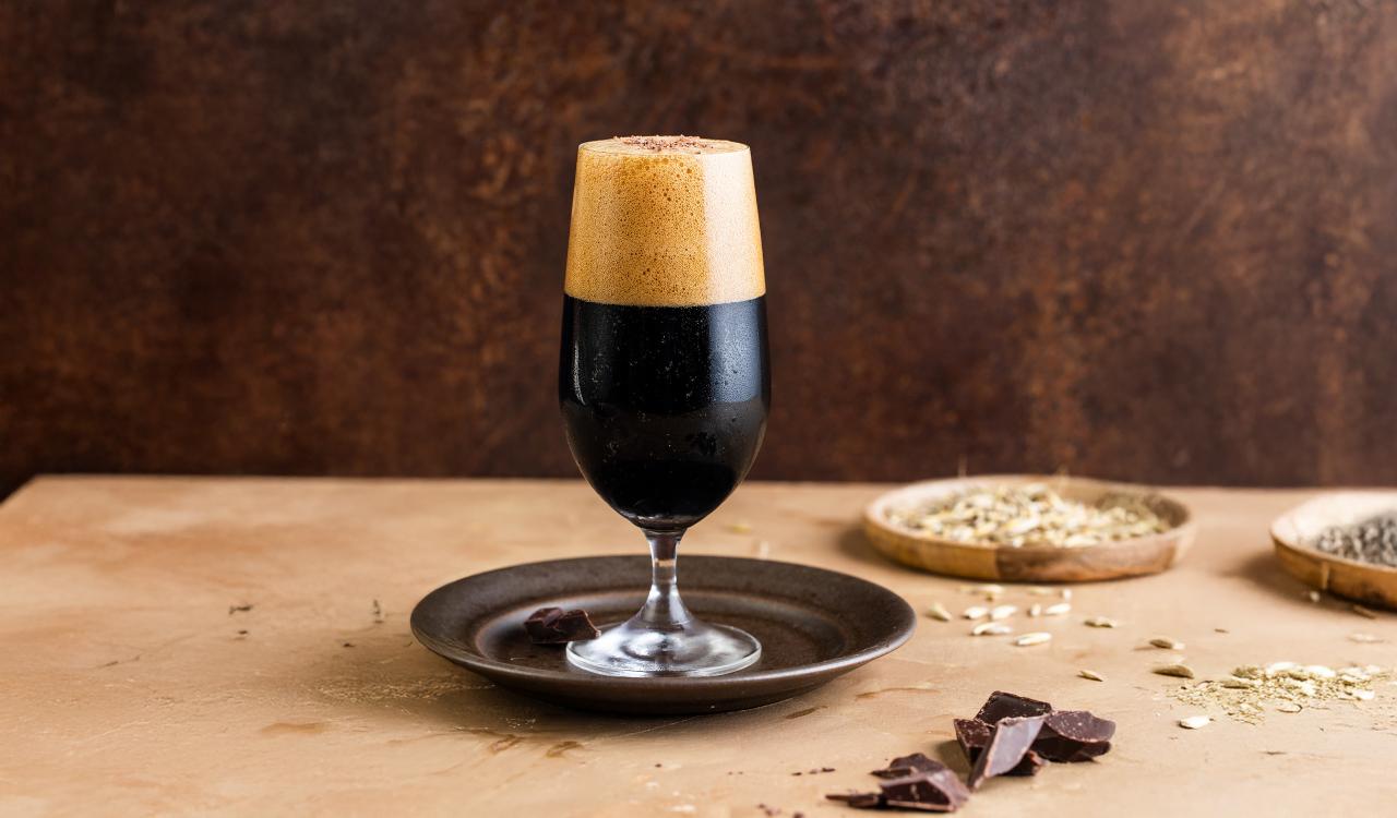 Glas mit dunklem Bier und cremigem Schaum auf einem dunklen Untersetzer vor braunem Hintergrund mit Schokoladenstücken und Schalen mit Körnern