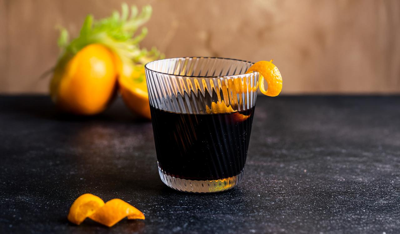 Glas mit dunklem Cocktail und Zitronenzeste auf dunklem Untergrund mit Zitronen im Hintergrund
