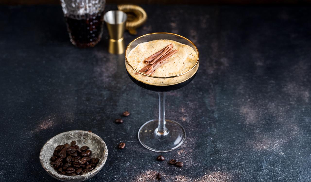 Cocktailglas mit schaumigem Espresso Martini und Zimtstangen auf dunklem Untergrund mit Kaffeebohnen und Messbecher im Hintergrund