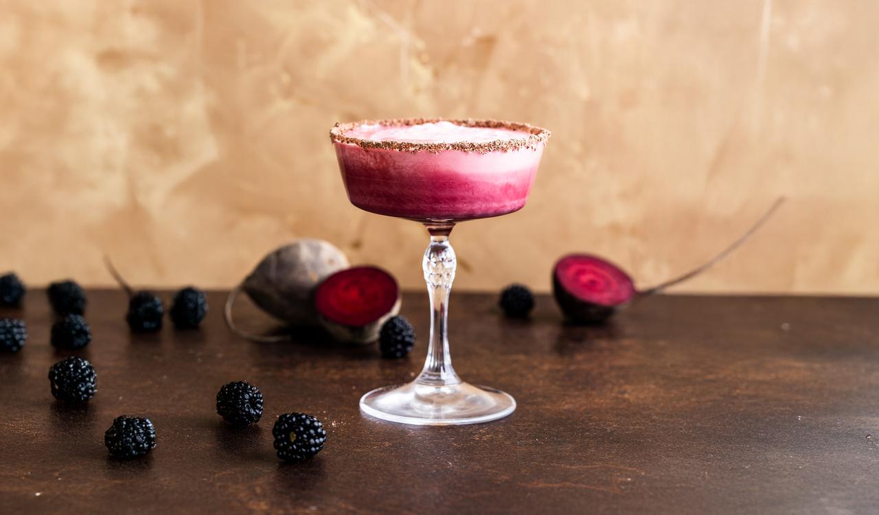 Cocktailglas mit rosa Schaum und Zuckerrand auf dunklem Untergrund umgeben von Brombeeren und halbierten Rote-Bete-Knollen