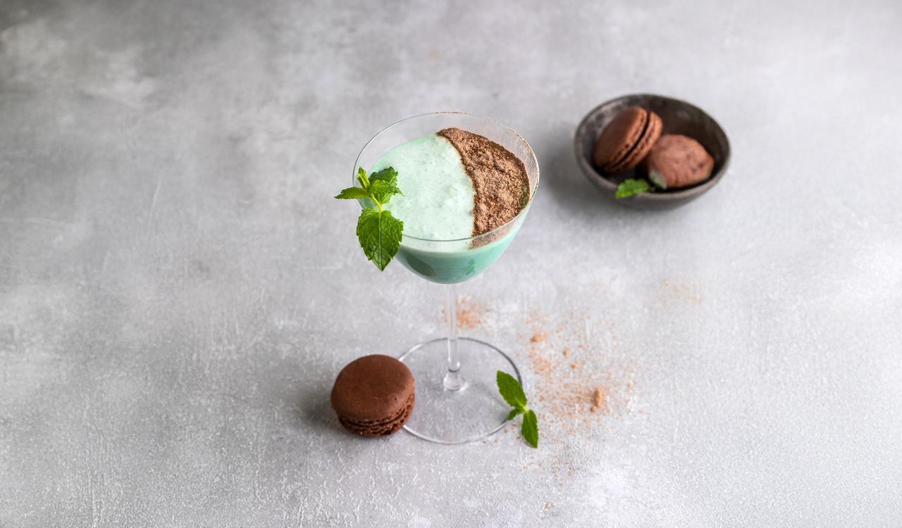 Cocktailglas mit grünem Getränk, mit Kakaopulver und Minzblatt garniert, daneben zwei braune Macarons auf grauem Untergrund