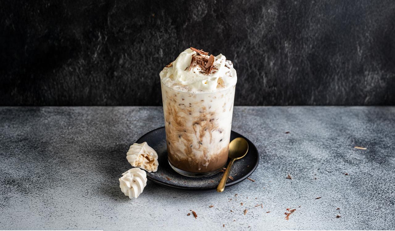 Glas mit Eiskaffeegetränk, Sahnehaube und Schokoraspeln auf dunklem Unterteller mit goldfarbenem Löffel und zwei kleinen Baiserstücken auf grauem Untergrund